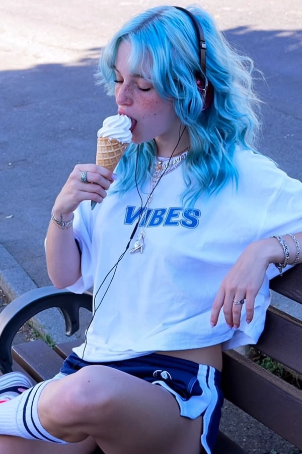 A spontaneous candid capture of a young woman with vibrant pastel blue hair styled in loose tousled waves, wearing a loose white crop top emblazoned with the word “VIBES,” paired with sporty-chic casualwear. She sits relaxed on a wooden park bench eating an ice cream cone, headphones in place, exuding an unbothered, serene expression. The ambient outdoor setting features soft, natural daylight subtly tinted with muted blue hues, creating a low contrast scene with distinctly cool-toned shadow areas in soft lavender-gray and blue. Textural details include visible skin freckles, gently defined loose hair strands in vivid pastel blue tones, fabric wrinkles on the cotton crop top, and subtle accessories barely visible. Composition employs an intuitively tilted, slightly off-center framing with casual cropping reminiscent of mid-2010s informal smartphone snapshots. The image embraces intentionally muted saturation, softened contrast, and cool-toned shadows typical of mid-2010s candid Tumblr-style photography.