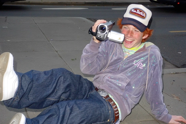 Caught mid-laugh under a soft sunlit street glow, a freckled redhead boy leans casually, gripping a chunky handheld camcorder as a lens flare gleams playfully across his face. He sports a glitter-speckled velour hoodie zipped over a layered pastel tank, baggy low-rise jeans cinched with a decorative belt, and a tilted Von Dutch trucker hat that’s slightly pixelated by the gentle CCD grain. Chunky white Nike Air Force 1s ground the laid-back Y2K vibe. The photo quality is low-resolution and somewhat grainy, with blurry edges and a subtle flash shine that wraps the moment in authentic early-2000s casual charm. The off-kilter framing feels spontaneous, like a chill streetboard snapshot made just for fun. —casual candid early-2000s Y2K snapshot, grainy low-res softness