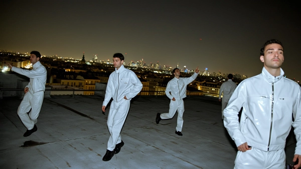 A spontaneous, surreal iPhone snapshot depicting a man duplicated multiple times on a wide urban rooftop at night, dressed identically in a fully white vinyl outfit with a high collar and detailed zippers. Some figures stand still in ambient city light casting soft reflections on the shiny material, while others are mid-step or reaching contemplatively toward the skyline. The figures drift in and out of shadow, with varied scales and positions creating layered depth. The natural glow of distant city lights subtly illuminates the textured vinyl fabric and the man's relaxed but focused expressions. Framing includes casual angles with slight cropping, enhancing an authentic, candid feel infused with a dreamlike repetition and surreal scale.