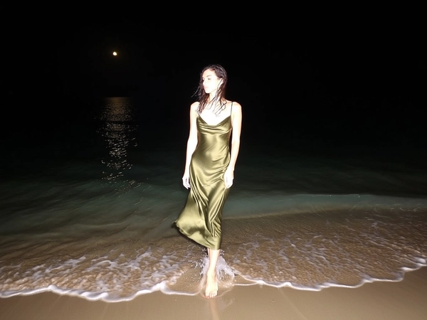 Her silhouette appears sharply against the inky blackness as a sudden flash rings out, catching the subtle shimmer of an olive-toned satin dress hugging her frame, its thin straps resting lightly on poised shoulders. The gentle tide licks the sandy shore, rippling softly around her bare feet as a faint lunar gleam dances on the water's surface behind her. Strands of damp hair cling tenderly, teased by a cool breeze, while the depths of the night stretch wide as negative space, framing her slightly off-center with nuanced blur where the hem sways. Calm and composed, she stands with relaxed arms, embodying quiet magnetism in this flash-lit elegant beach snapshot, captured on iPhone.