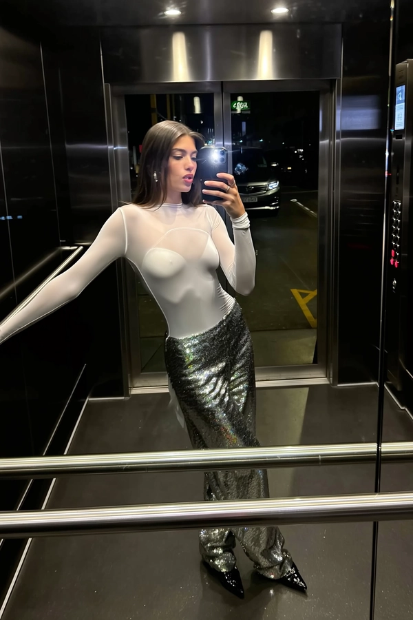 Cool flickers of stark white LED dance off the translucent overlay of her long-sleeve top, the sheer fabric whispering softness against the shimmer of low-slung sequin trousers that catch the faint pulse of light below. Black stilettos glint like onyx shards on the glass floor, sharp contrasts gliding beneath the suspended cab, soaring above the shadowy sprawl of an underground garage. Her silhouette is caught mid-lean, one arm blending seamlessly with the glass wall’s metallic framing, phone lowered to chest level capturing the crisp weave of satin and the flicker of reflected finger smudges. She gazes at the screen, unbothered, lips set in neutral rhythm, as the cool urban night hums just beyond the elevator’s glass, the occasional electric flicker softening the edges of this quiet moment—elevator mirror capture, urban chic on iPhone