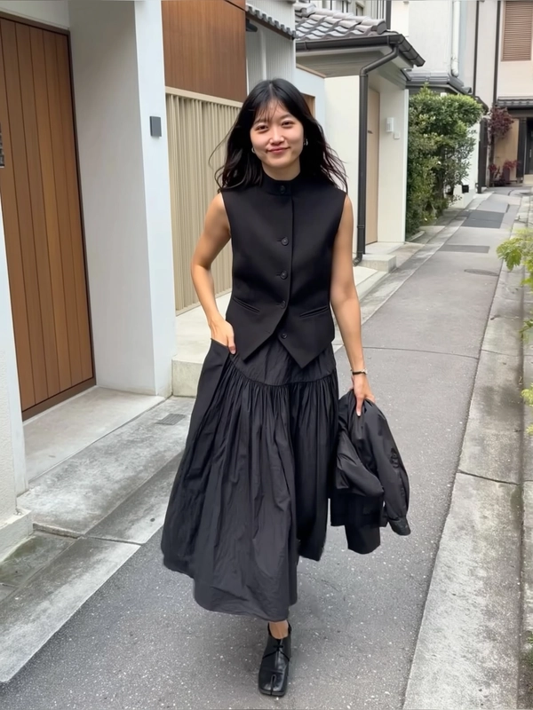 A spontaneously candid iPhone photograph capturing a woman walking toward the camera down an empty pedestrian path set in a quiet residential neighborhood characterized by a subtle row of minimalistic doors and muted neutral tones. She wears a sleeveless structured black vest paired with a crisply creased full black skirt embodying minimalist Japandi elegance, complemented by black platform shoes. One hand is casually tucked into her pocket while the other holds a folded lightweight jacket with natural ease. Soft diffused outdoor daylight gently illuminates the scene, revealing tactile fabric textures in her garments, subtle natural skin details, and loose hair strands. The composition features a slight casual tilt and off-center framing, reinforcing the authentic spontaneous feel of an unposed iPhone snapshot. The surrounding architecture incorporates clean lines and muted colors evoking Japandi aesthetics — subtle concrete paths, neutral wood-tone door frames, and sparse greenery lending warmth and simplicity to the atmosphere.