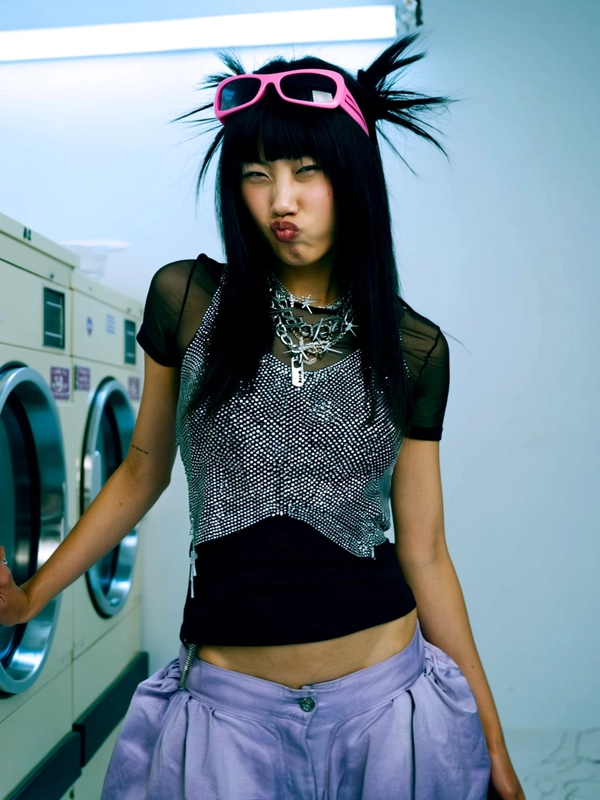 She stands in a medium shot before the softly blurred spinning drum of a laundromat machine, framed awkwardly as if caught mid-adjustment by a handheld digital cam. Her sheer rhinestone-studded top sparkles against a tight black tank underneath, paired with low-rise flared pants in an icy lavender tone that catch the dim neon glow from overhead. Her eyes are squinted sharply, lips pursed with calculated detachment; the steeliness in her gaze cuts through the sterile backdrop while her dark hair cascades loose with subtle gelled spikes, catching the cold studio light. Accessories include layered silver chain necklaces with punk spikes and a pair of oversized plastic pink sunglasses perched atop her head, accentuating the deliberate, unbothered attitude. The composition feels both casual and editorial—a perfect frozen moment of Y2K street-meets-studio cool. —casual candid early-2000s Y2K snapshot, grainy softness