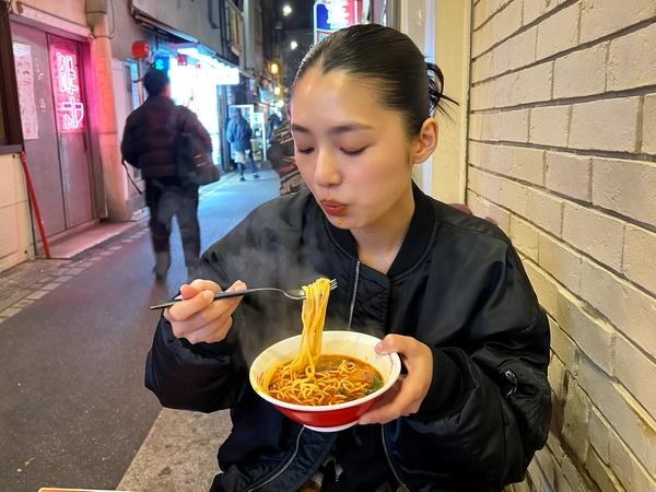 Amid the narrow glow of a bustling Tokyo alley, a young brunette woman sits perched on a low stool, her hair pulled back in a sleek bun that reveals a smooth, flawless complexion. She wears a softly tailored black bomber jacket, the matte fabric catching scattered neon reflections, while a delicate silver chain lays just against the curve of her neck. Her face is calm and measured, eyes half-lidded yet glassy with the faint sheen of tears, lips parted as she lifts a lacquered black spoon toward her mouth.

Steam curls in gentle wisps from the bowl of fiery ramen cradled in her hands, the glossy, chili-spiked broth shimmering beneath strands of bright, taut noodles. The precise movement of her fork twirling the tangled noodles glistens with droplets of spicy sauce, a tiny glint on her lip gloss catching just as she delicately slurps a mouthful. Her gaze remains steady, maintaining serene composure despite the subtle flush of heat rising in her cheeks.

The alley’s ambient light, a mix of cool blues and warm pinks from flickering neon signs, filters lightly through the mist and steam, suffusing the scene with a cinematic, late-night aura. Textured wall bricks and scattered wet pavement reflect the mixed hues while faint silhouettes of pedestrians pass by, providing a lively yet intimate urban backdrop.

Captured from a casual, arm’s-length angle just above waist level, the image holds a natural sense of immediacy and quiet focus. The tactile richness from glistening broth to softened hair strands invites the viewer into this flavorful, tranquil moment, perfectly resembling a candid iPhone photo.
